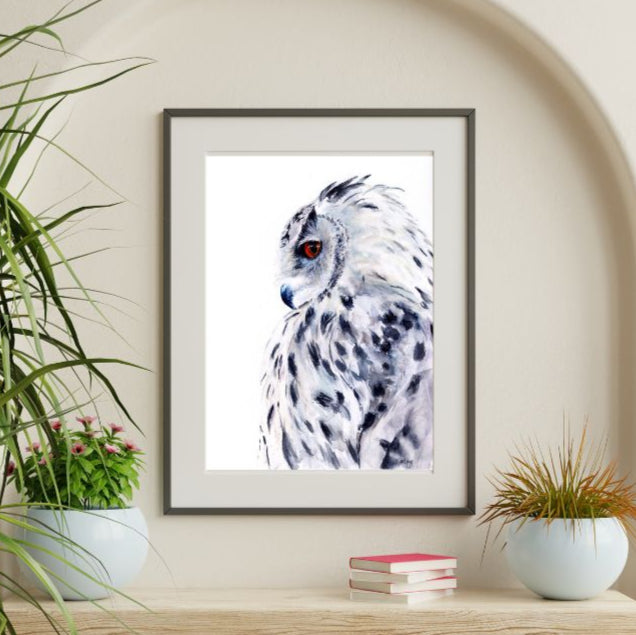 Framed artwork of a snowy owl on a wall above a wooden shelf with plants and books.